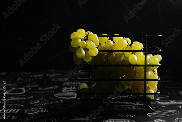 Fototapeta Kishmish grapes in a metal black grid stand, on a black background, in a low key, dark photo