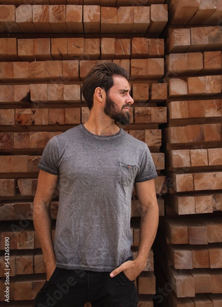 Fototapeta Caucasian bearded man in profile with hands in pockets. Stylish handsome young man dressed casual. Wood background.