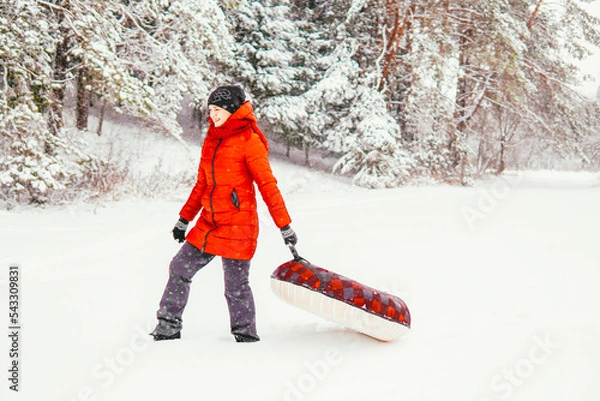 Fototapeta a girl in a red jacket holds a tubing and laughs in winter against the backdrop of a snowy forest