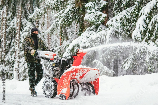 Fototapeta a man cleans the road from snow with a snowplow in the forest in winter against the backdrop of a snowy forest