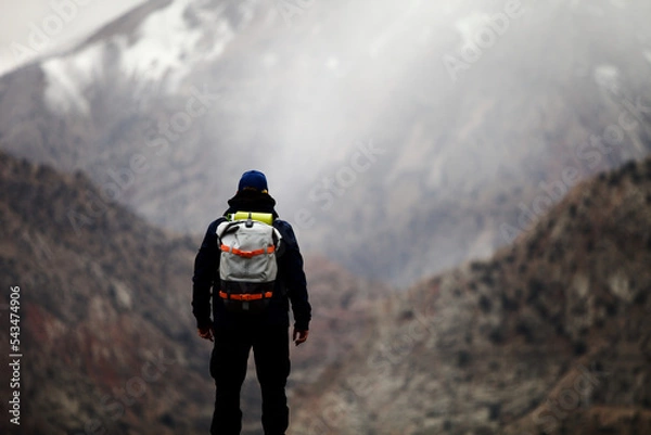 Fototapeta A man with backpack stands on a mountainside and looks into the distance at a mountain peak. A lone hiker travels high in the mountains on a cloudy day. Independent hike in Pamir mountains. 