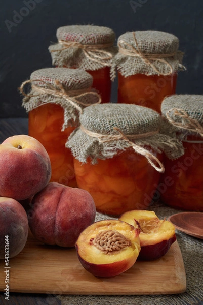 Fototapeta There are several fresh peaches on a cutting board, one of them cut in two. Jars of peach jam sit on a dark wooden table at the back.