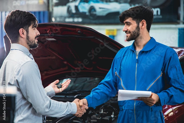 Fototapeta Man picking up car from garage a handsome male mechanic in a uniform returns the keys to the customer and shake hands with customers to thank.