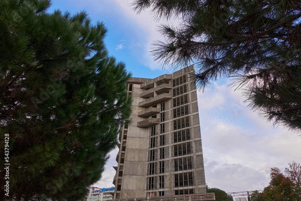 Fototapeta An apartment building under construction. Concrete structure with balconies and window openings. Pines ahead. Blue cloudy sky.