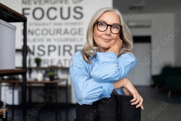 Obraz confident businesswoman with eyeglasses sitting with her arms folded comfortably on the back of a chair and looking at the camera, caucasian Middle aged older businesswoman at work Concept