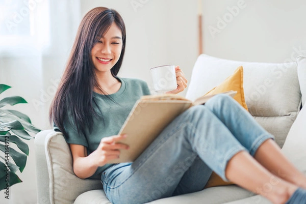 Fototapeta Young asian woman sitting on modern sofa in front of window relaxing in her living room reading book and drinking coffee or tea at home