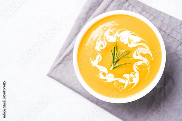 Fototapeta Thick butternut squash bisque in white bowl garnished with heavy cream and rosemary over white table. Bowl of pumpkin cream soup. View overhead. Minimal style. Selective focus