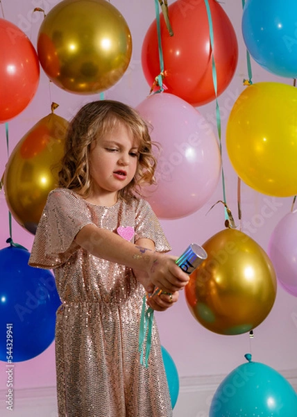 Fototapeta Young girl in a sparkly party outfit blowing up the confetti сannon. Balloons on the background.
