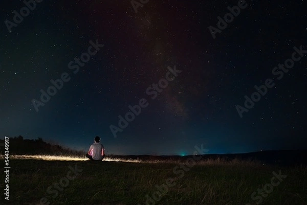 Fototapeta Beautiful night sky with stars and silhouette of a standing alone man on the mountain with a lantern. Blue milky way and man on the hill. Background with galaxy and silhouette of a man. 