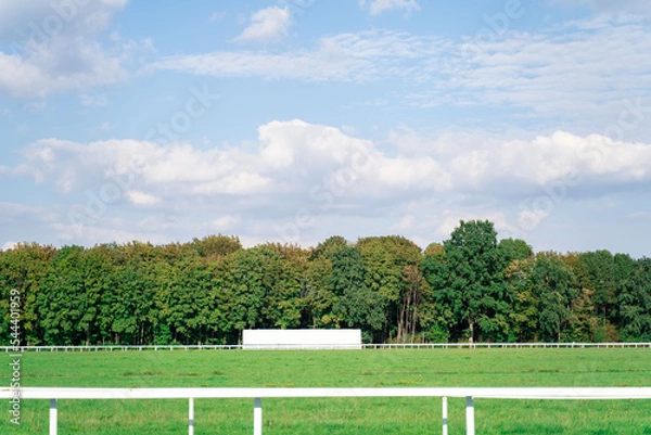 Fototapeta Empty high-speed track with partitions at hippodrome for horse racing surrounded by dense green forest with trees and cloudy blue sky on background. Gambling, betting, weekend activity, holiday events