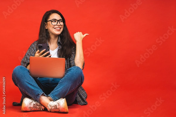 Fototapeta Business concept. Portrait of happy smiling brunette woman in casual sitting on floor in lotus pose and holding laptop isolated over red background. Using mobile phone.