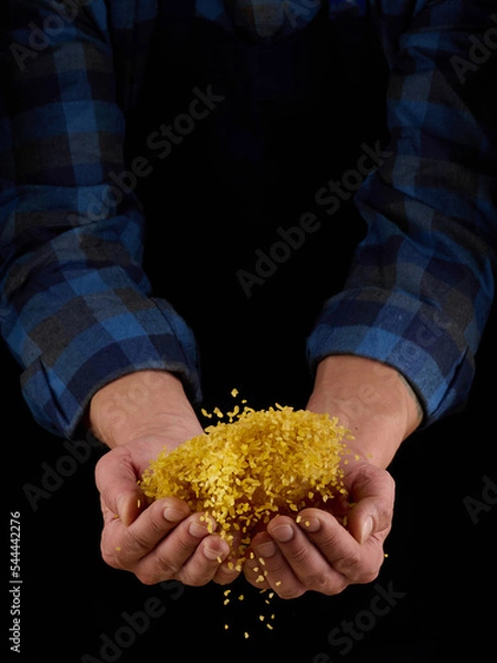 Obraz Bulgur grains in hands on a dark background. Hands of man pour grain of bulgur. Close-up