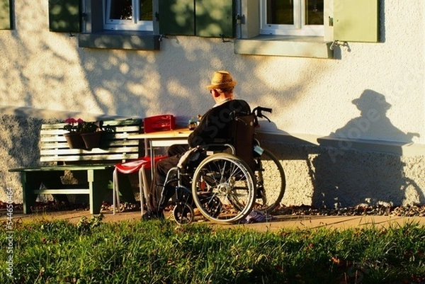 Fototapeta Alter Mann im Rollstuhl sitzt vor seinem Haus in der Abendsonne