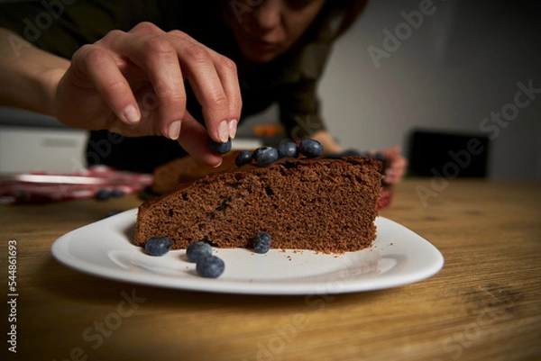 Fototapeta Woman with a passion for cooking decorates a slice of handmade chocolate cake in her home kitchen with blackberries and red currants.