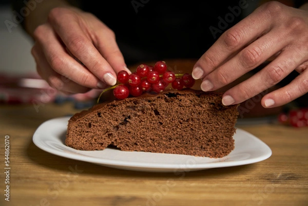 Fototapeta Woman with a passion for cooking decorates a slice of handmade chocolate cake in her home kitchen with blackberries and red currants.