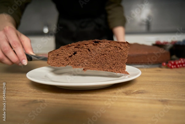Fototapeta Woman with a passion for cooking decorates a slice of handmade chocolate cake in her home kitchen with blackberries and red currants.