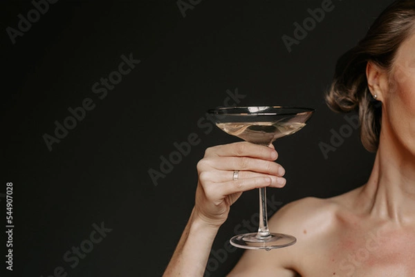 Fototapeta Half the face of woman with open shoulders, holding glass of champagne on background of a black wall in isolated studio