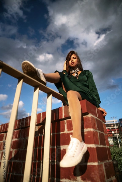 Obraz Street style fashion portrait of a cute young brown haired girl in a green dress under the cloudy sky. Attractive slim woman sits on the fence and poses in the light of the sun.