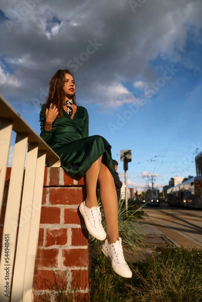 Obraz Street style fashion portrait of a cute young brown haired girl in a green dress under the cloudy sky. Attractive slim woman sits on the fence and poses in the light of the sun.
