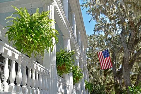 Obraz The columns and railings of an old southern home are graced with baskets of ferns and the American flag.