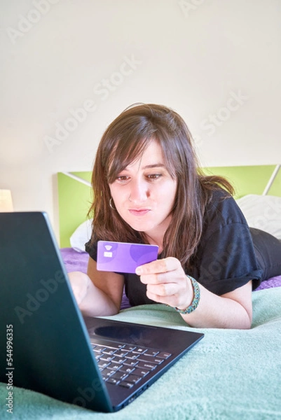 Fototapeta Vertical closeup of the woman looking at the credit card lying on the bed in front of the laptop.