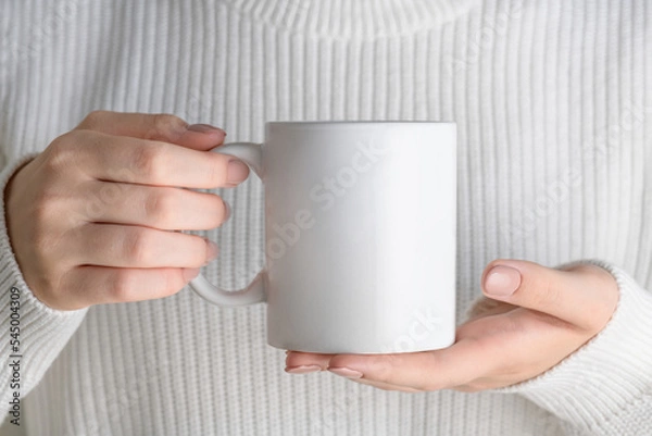 Obraz Female hand holding white mug mockup with blank copy space for your advertising text message or promotional content. Girl in blue shirt holding white porcelain coffee mug mock up