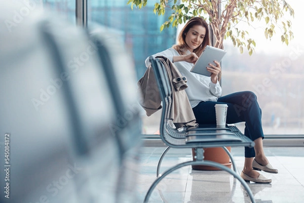 Fototapeta Young smiling woman using a tablet and waiting for a flight in the departure hall at the airport.