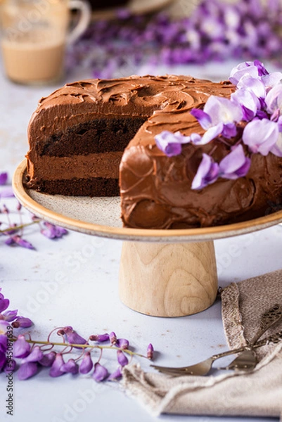 Fototapeta Sliced chocolate brownie cake on a cake stand decorated with fresh purple flowers, frosted and iced with cocoa ganache