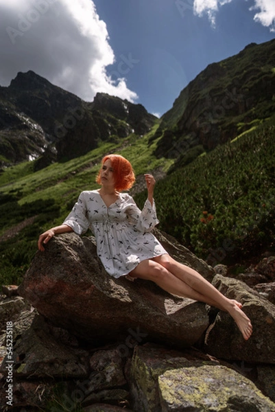 Obraz Elegant sexy lady with red hair in a white dress sits on the rock among spruce in the mountains and relaxes under sun rays. Fashion natural portrait as wallpaper, magazine design or advertising cover.