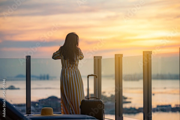 Obraz Beautiful woman in dress with suitcase on vacation by tropical beach, standing on the glass balcony enjoy fresha air with ocean view. Golden sunset sky on the beach, Beautiful cloudscape over the sea.