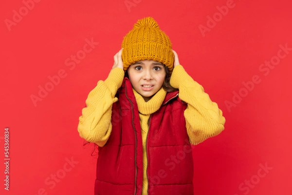 Fototapeta kid in knitted winter hat and down vest feel cold on red background, fashion
