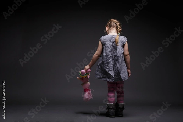 Fototapeta Rear image of a little sad girl with doll, toddler, offended by someone, being in bad mood, over dark grey background.