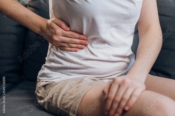 Fototapeta Close up of young female with fatty liver touches right side with hand, suffering from abdominal pain sit on grey couch. Pain in right side, appendix, gallstones and gynecological diseases concept