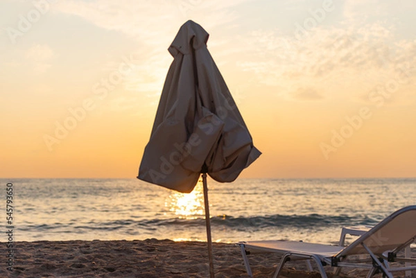Obraz Sun lounger and umbrella on the background of the sunset sea. Folded beach umbrella covers the sun.
