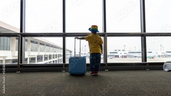 Fototapeta Child with suitcase at airport terminal waiting for departure looking out the window. Child and suitcase at the airport, indoors and waiting for going to travel.	