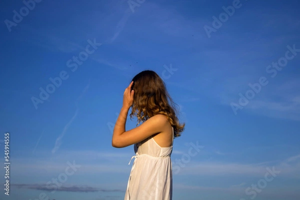 Fototapeta a girl with long curly hair in a white sundress stands with her back against the blue summer sky