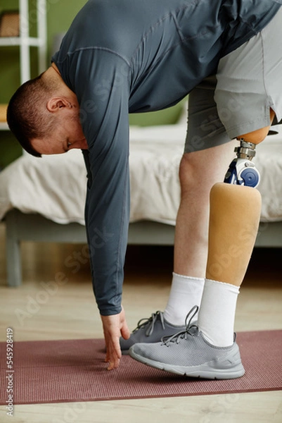 Fototapeta Vertical portrait of man with prosthetic leg working out at home and doing stretching exercises