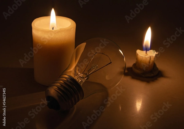 Fototapeta Switched off light or not glowing light bulb near a burning candles in total darkness during power cut. Blackout city, electricity off, load shedding, energy crisis or power outage, concept image.