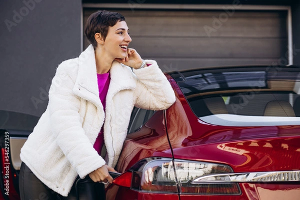 Fototapeta Woman charging electro car with charging pistol by the house