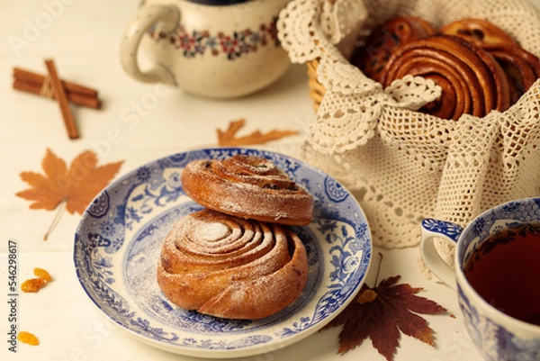 Fototapeta Tea party, break. Homemade cinnamon and raisin buns on vintage plates, a teapot and a wicker basket with buns in the background.
