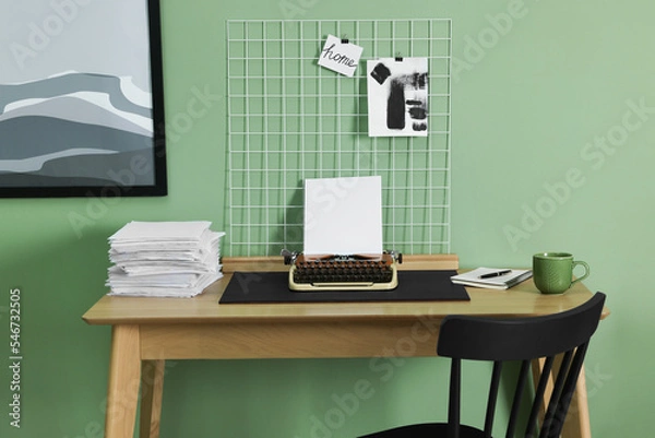 Fototapeta Typewriter, stack of papers and mood board on wooden table near pale green wall. Writer's workplace