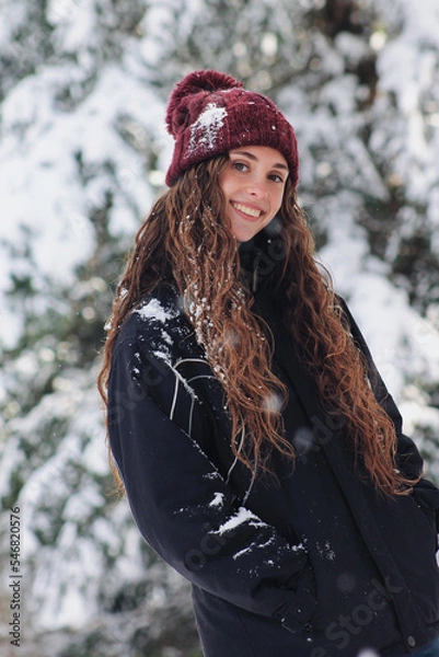 Fototapeta portrait of a pretty happy woman on a snowy day in winter, with her hands in her pockets. Woman bundled up in a snowflake hat and anorak. Cold and snowy winter landscape. Enjoying winter and snow.