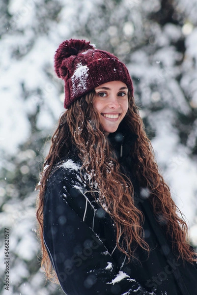 Fototapeta portrait of a happy woman on a snowy day in winter. Woman bundled up in a hat and anorak. Snowy cold forest. Snow flakes. Enjoying winter and snow. Caucasian woman with long hair and hat.