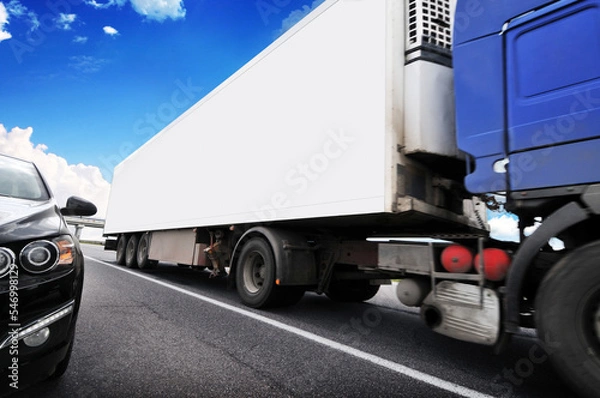 Fototapeta Close-up of a blue truck with a white trailer on a highway with other car against a sky with clouds