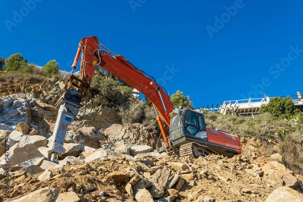 Fototapeta Heavy organge excavator with shovel standing on hill with rocks