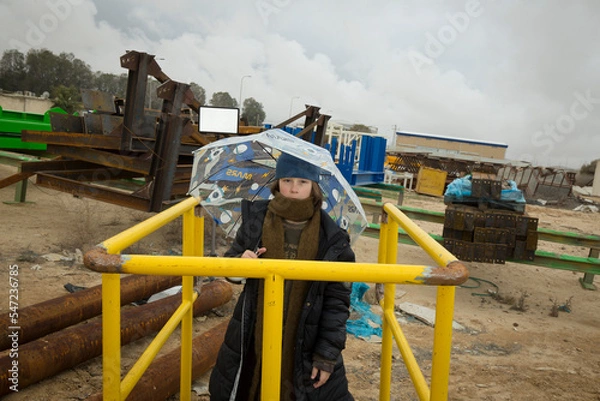 Obraz cool stylish boy in a coat and hat with a scarf posing plays and walks through pipes with an umbrella in an industrial area among green yellow blue metal structures against a cloudy sky on a rainy day