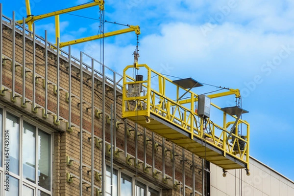 Fototapeta Construction hoist on the facade of the building. House cladding work. Empty facade elevator on cables.