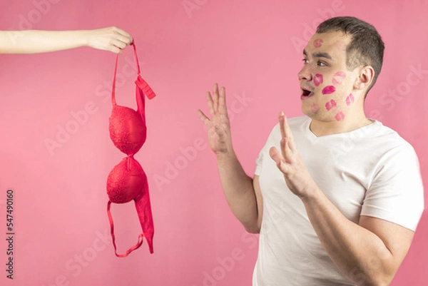 Fototapeta A woman's hand with a red bra and a man with traces of a kiss on his face in anticipation of sex, pink background. The concept of sex,sexual relations,love,sexual foreplay