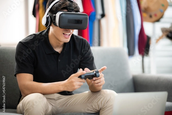 Fototapeta Young Asian male wearing headphones playing video games using virtual reality technology and hand console while sitting and relaxing at home during weekend while making facial expressions