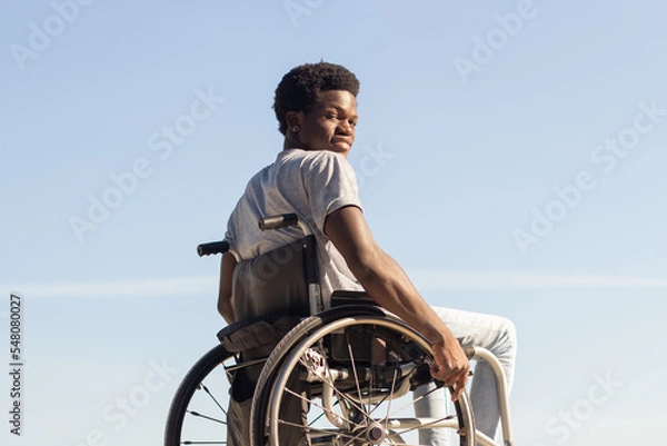 Fototapeta Black man in wheelchair enjoying sunny day in park, looking back and smiling at camera over the shoulder. Young African American guy with disability having fun outdoors. Disability, motivation concept